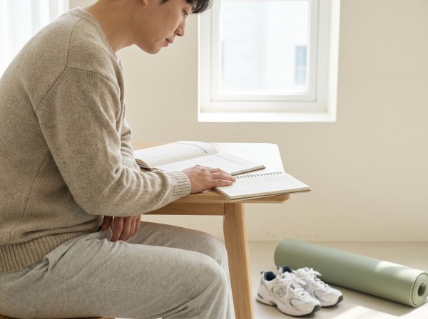밝고 차분한 집에서 편안한 일상복을 입은 성인이 달력과 운동 계획 노트를 살펴보는 자연스러운 사진. 옆에 운동화와 말린 운동 매트를 배치하고 상 - 수완지구골반필러 | 미미썸의원 광주첨단점